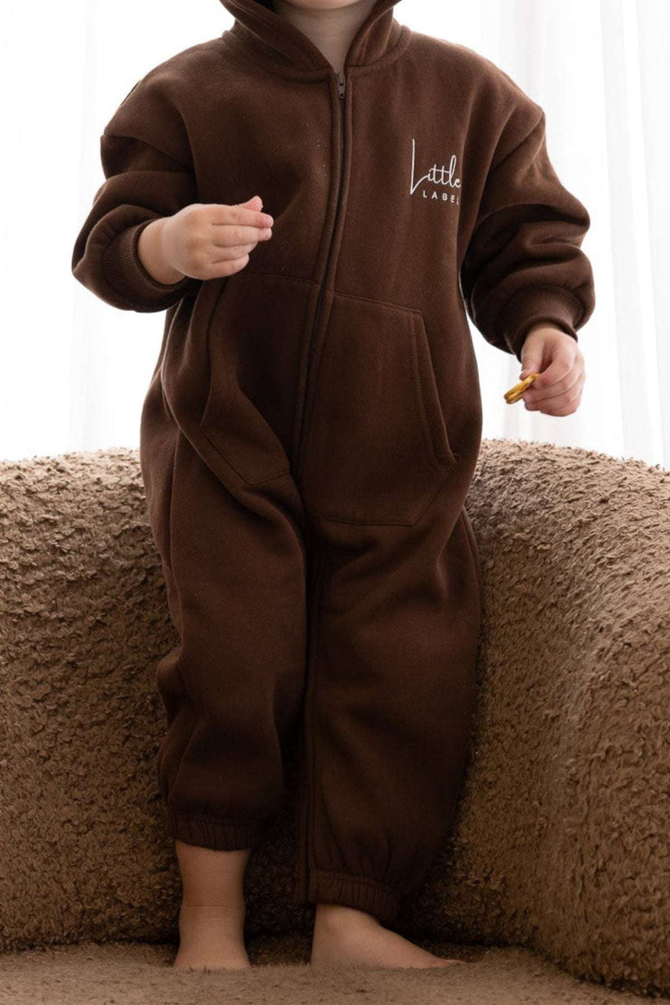 Child wearing a brown onesie with a logo, sitting on a textured surface.