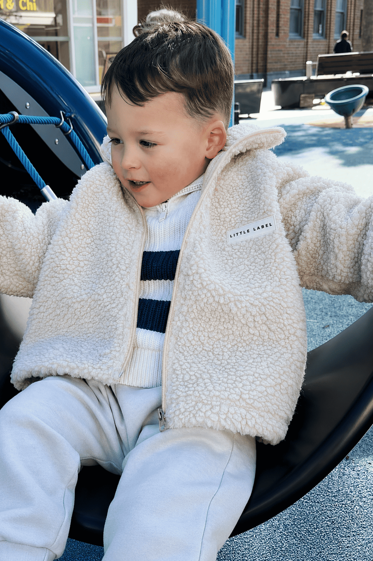 Child wearing a beige fleece jacket sitting on playground equipment | Ryan is 100cm tall and wears size 3Y.