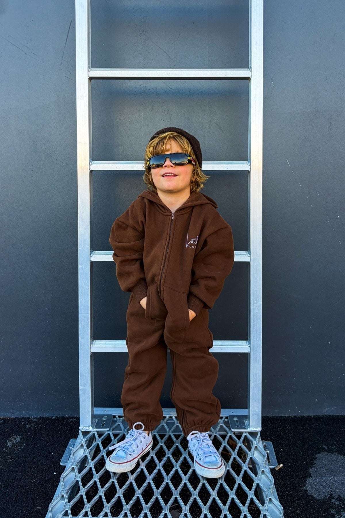 Child wearing a brown onesie standing on a metal ladder against a dark blue wall. | Luke is 3'2
