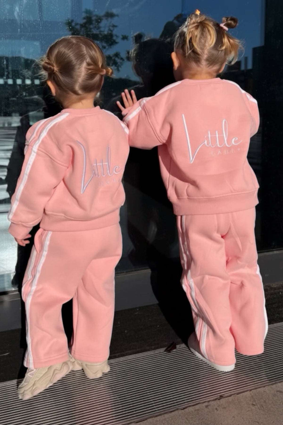 Two children in matching pink outfits with 'Little' on the back, standing close together.