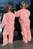 Two children in matching pink outfits with 'Little' on the back, standing close together.