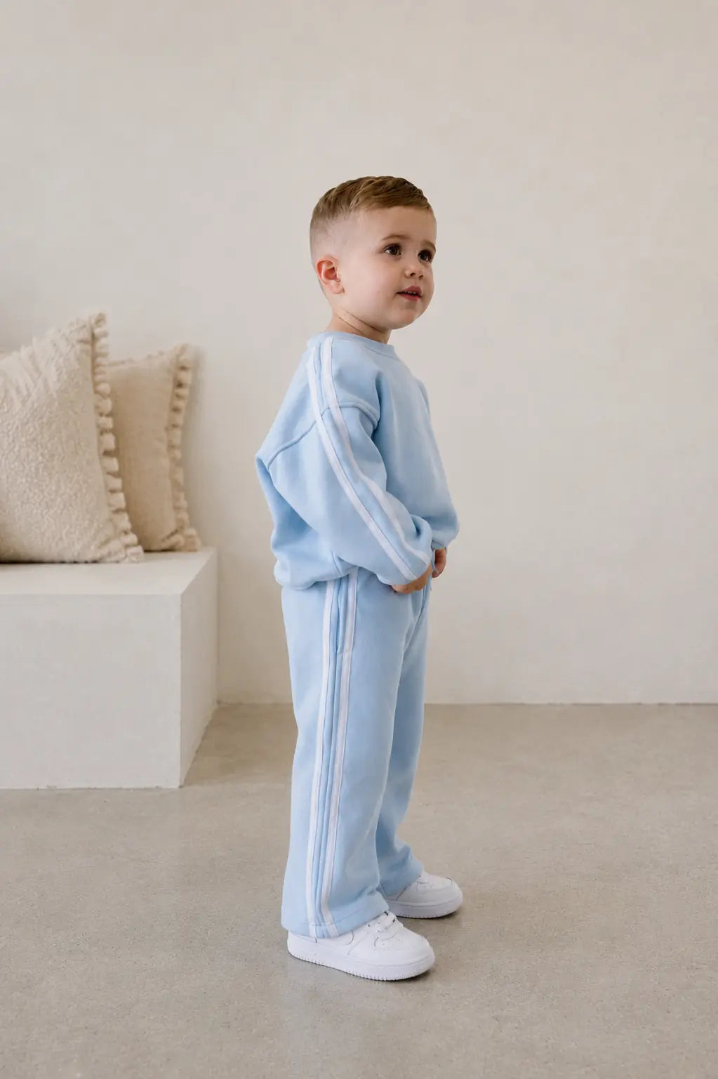 Child wearing a light blue tracksuit standing in a minimalistic room with beige pillows. | Alex is 2'6