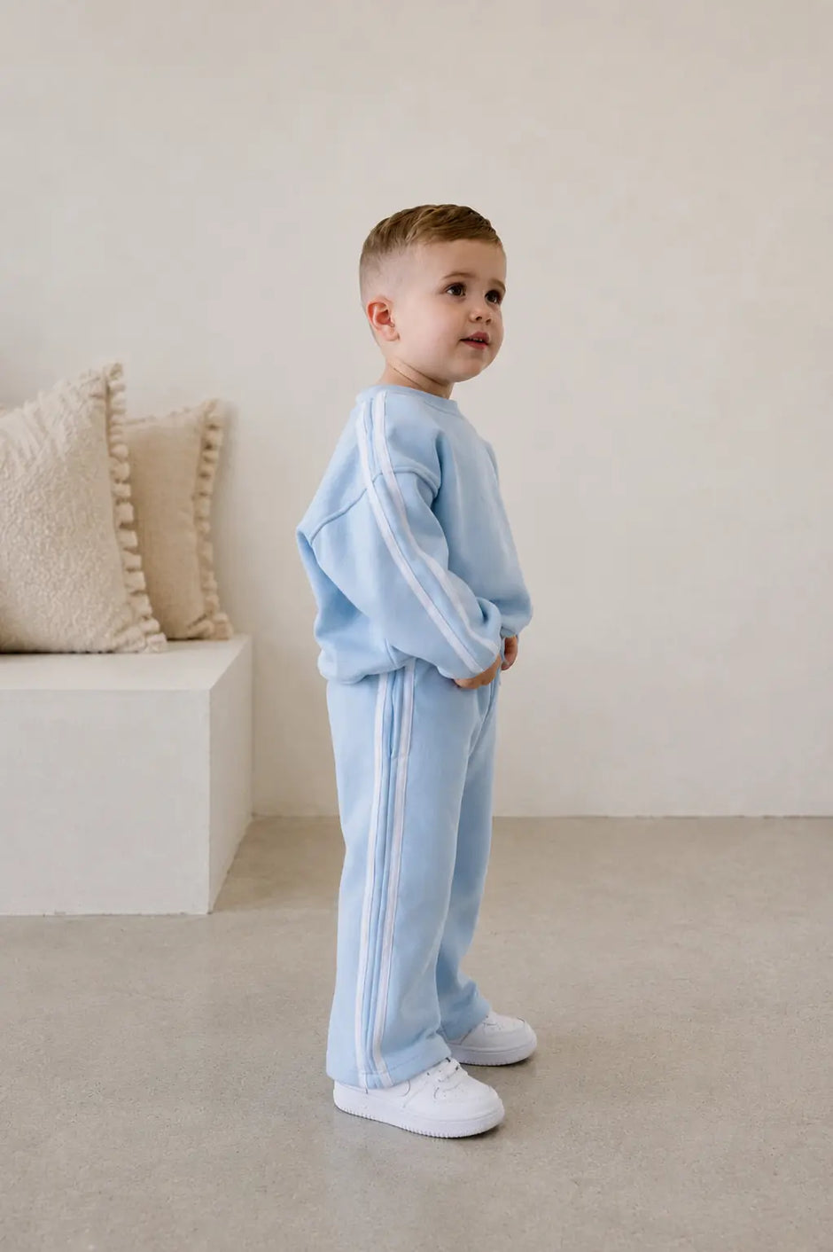 Child wearing a light blue tracksuit standing in a minimalistic room with beige pillows.