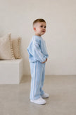 Child wearing a light blue tracksuit standing in a minimalistic room with beige pillows. | Alex is 2'6