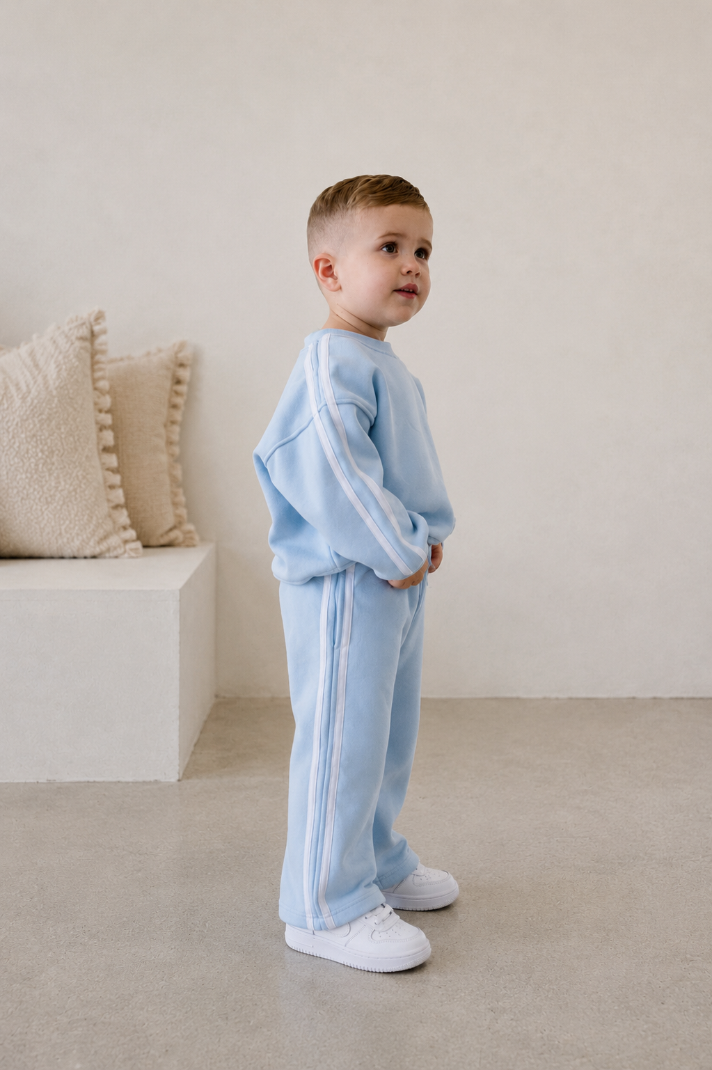 Child wearing a light blue tracksuit standing in a minimalistic room with beige pillows. | Alex is 2'6