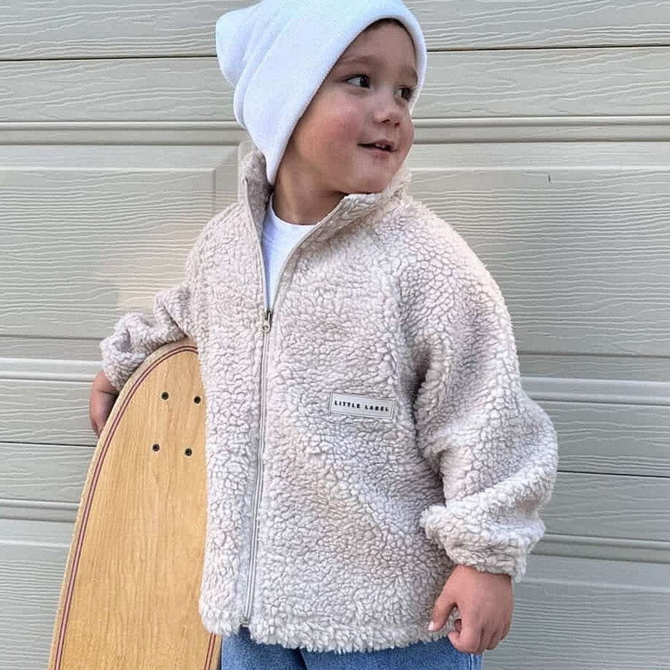 Child wearing a beige fleece jacket and white beanie holding a skateboard against a wooden background