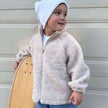 Child wearing a beige fleece jacket and white beanie holding a skateboard against a wooden background | Ryan is 3'3