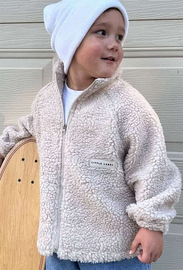 Child wearing a beige fleece jacket and white beanie holding a skateboard against a wooden background | Ryan is 3'3" (100cm), wears size 3Y