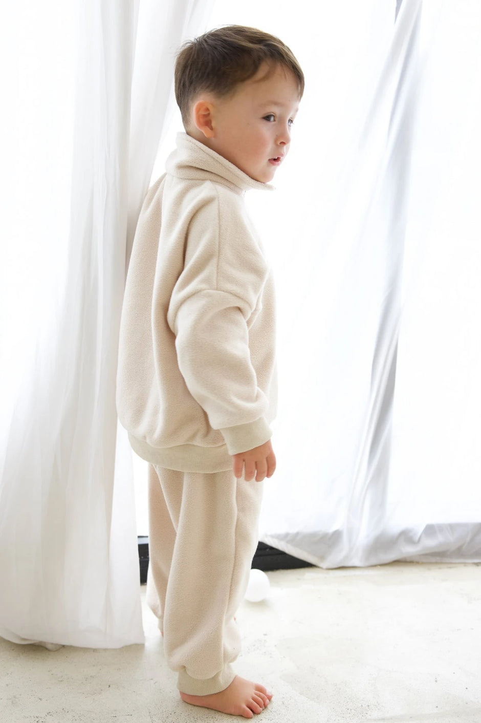 Child wearing a cream-colored outfit standing in front of white curtains.