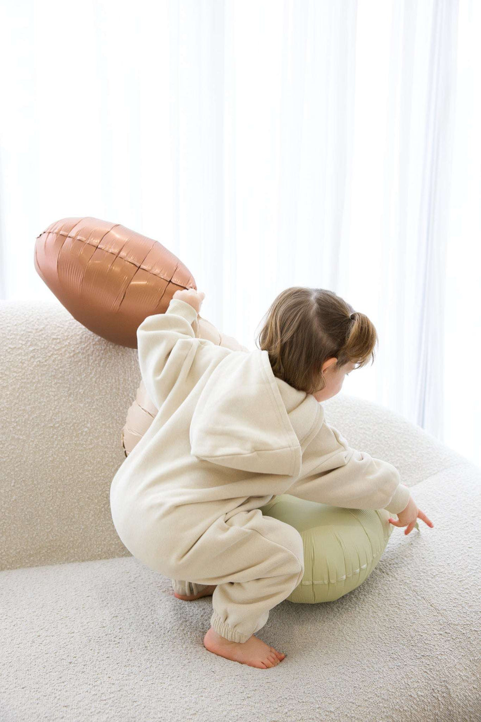 Child in a white onesie playing with a football on a light-colored couch