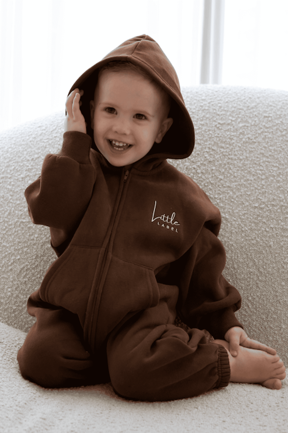 Child wearing a brown onesie with a hood, sitting on a light-colored couch.